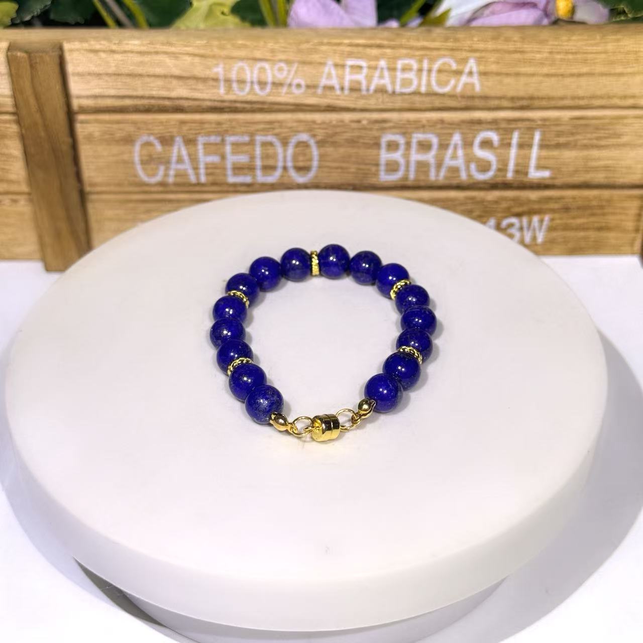 Classic minimalist natural 8mm Afghan Lapis Lazuli bead bracelet with delicate gold spacer wheels and a polished gold magnetic clasp, displayed on a clean white round jewelry riser against a wooden box background.